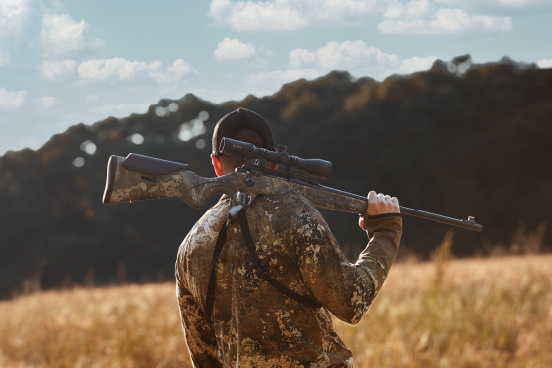 kfi monza - Popular Outdoorsman Person in camouflage clothing carries a rifle over their shoulders in an open field with tall grass and a treeline in the background.