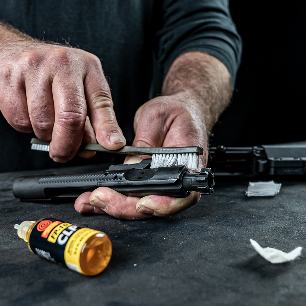 Otis cleaning AR 15 bolt - Popular Outdoorsman Close-up of hands cleaning a pistol slide with a brush, oil bottle nearby on a work surface.