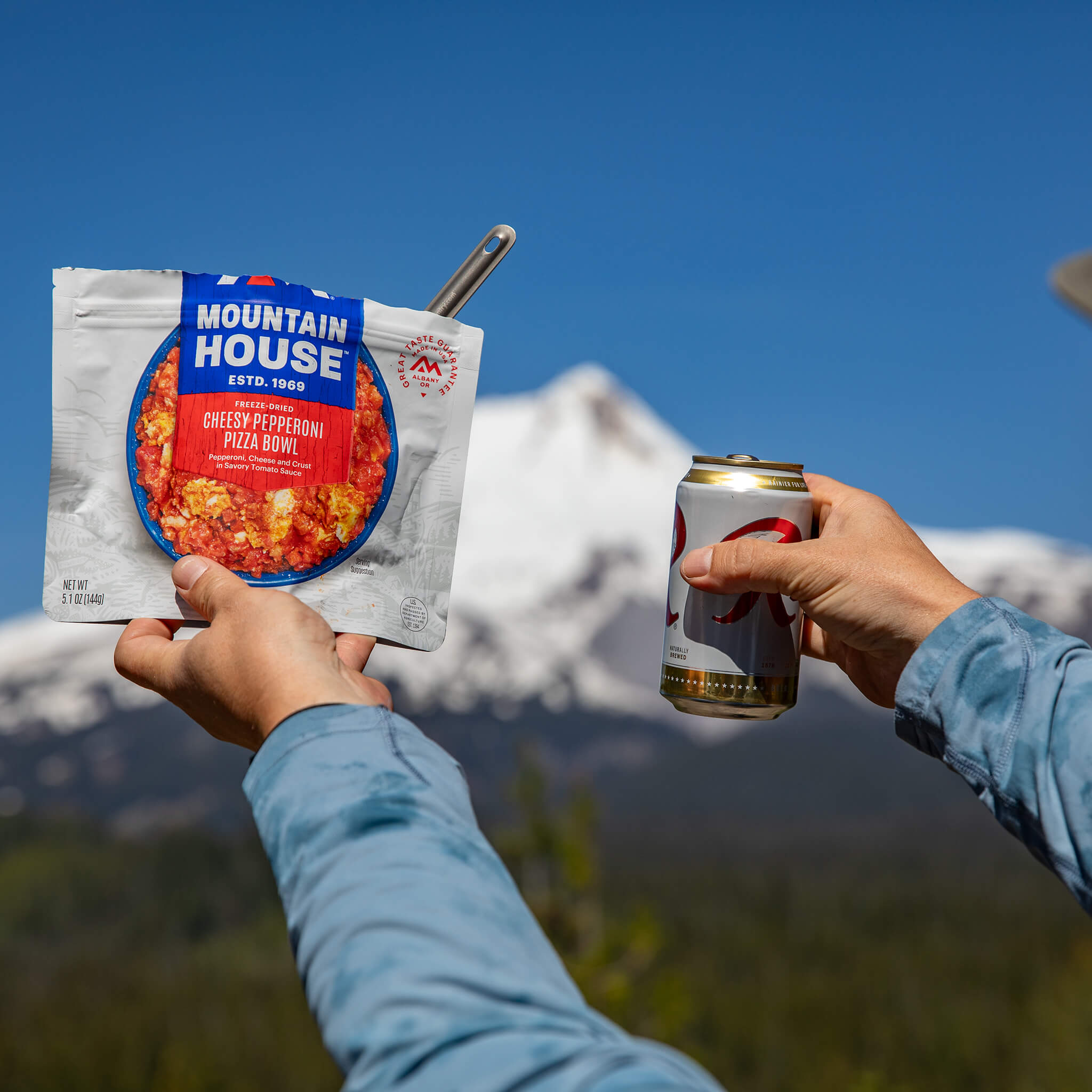 pizza-bowl-and-raineer-lifestyle - Popular Outdoorsman Two people hold a Mountain House meal pouch and a canned drink against a blue sky and mountain backdrop.