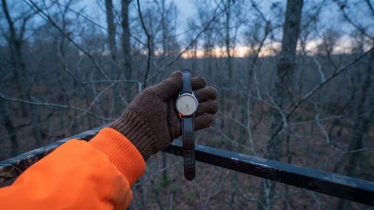 Legal shooting hours check in a tree stand at dawn: a gloved hand holds a wristwatch beside a blaze orange sleeve